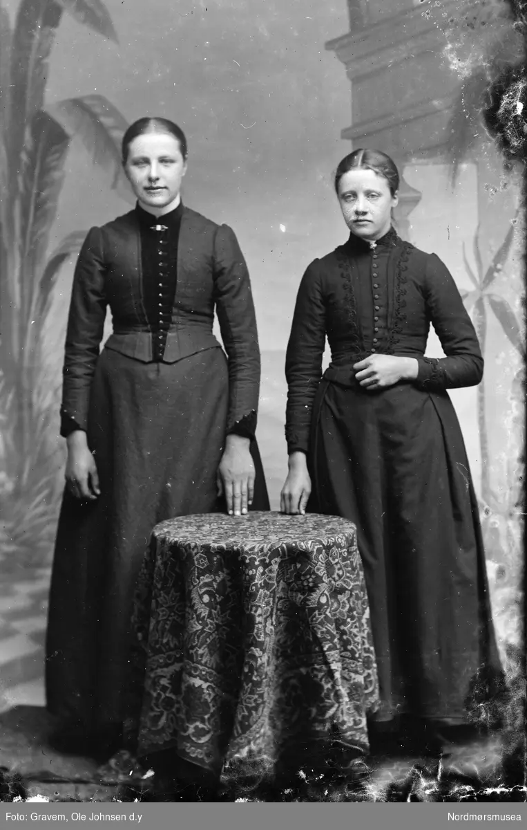 Portrett i helfigur av to kvinner. Kvinnene står bak et bord og har på seg kjoler.