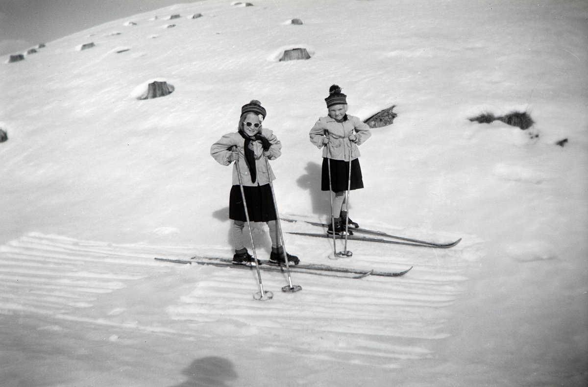 To jenter på ski fra Hol vestre i Vang til Nordre Arneberg i Romedal slutten av mars. Ikledd skjørt i anledning at de er på tur til en bursdag. Her over et hoggstfelt.  Jentene er Ragnhild Glærum (f: 1945) og Eli Vesaas (f: Glærum 1947). Fotograf Andrea Glærum (f: Gjestvang 1914)