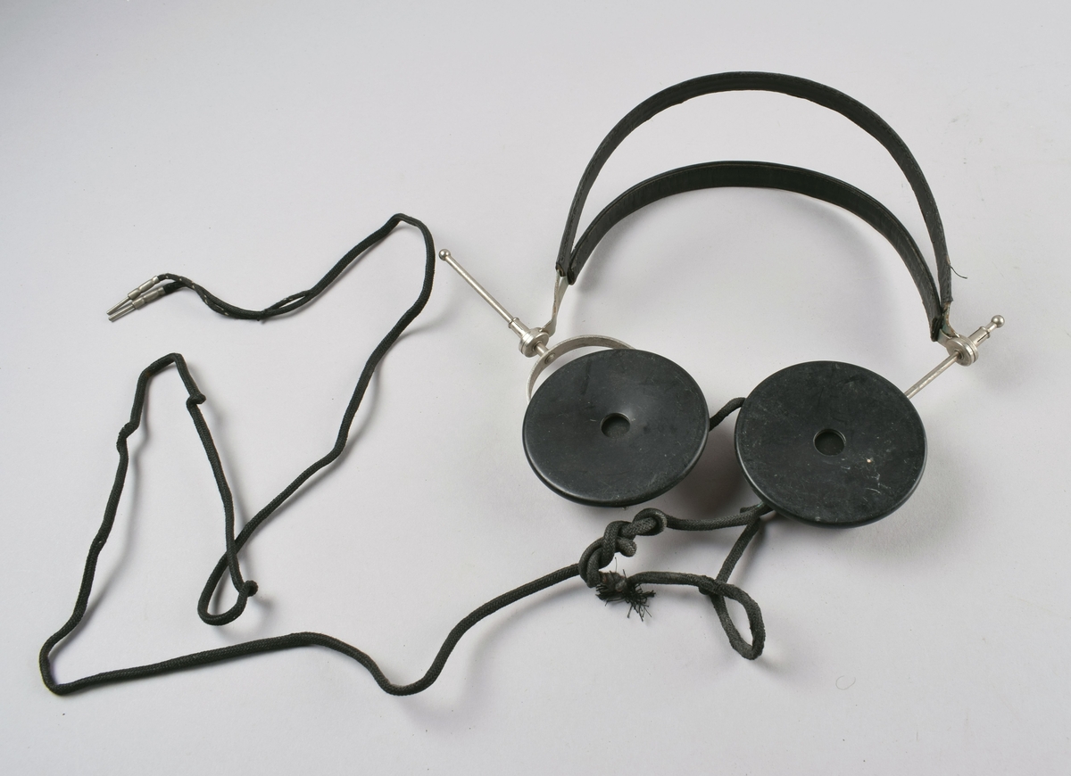 Høretelefoner forbundet med dobbel hodebøyle i metall trukket med skinn. Ledning som splittes i to plugger nederst. Hører til krystallapparat.