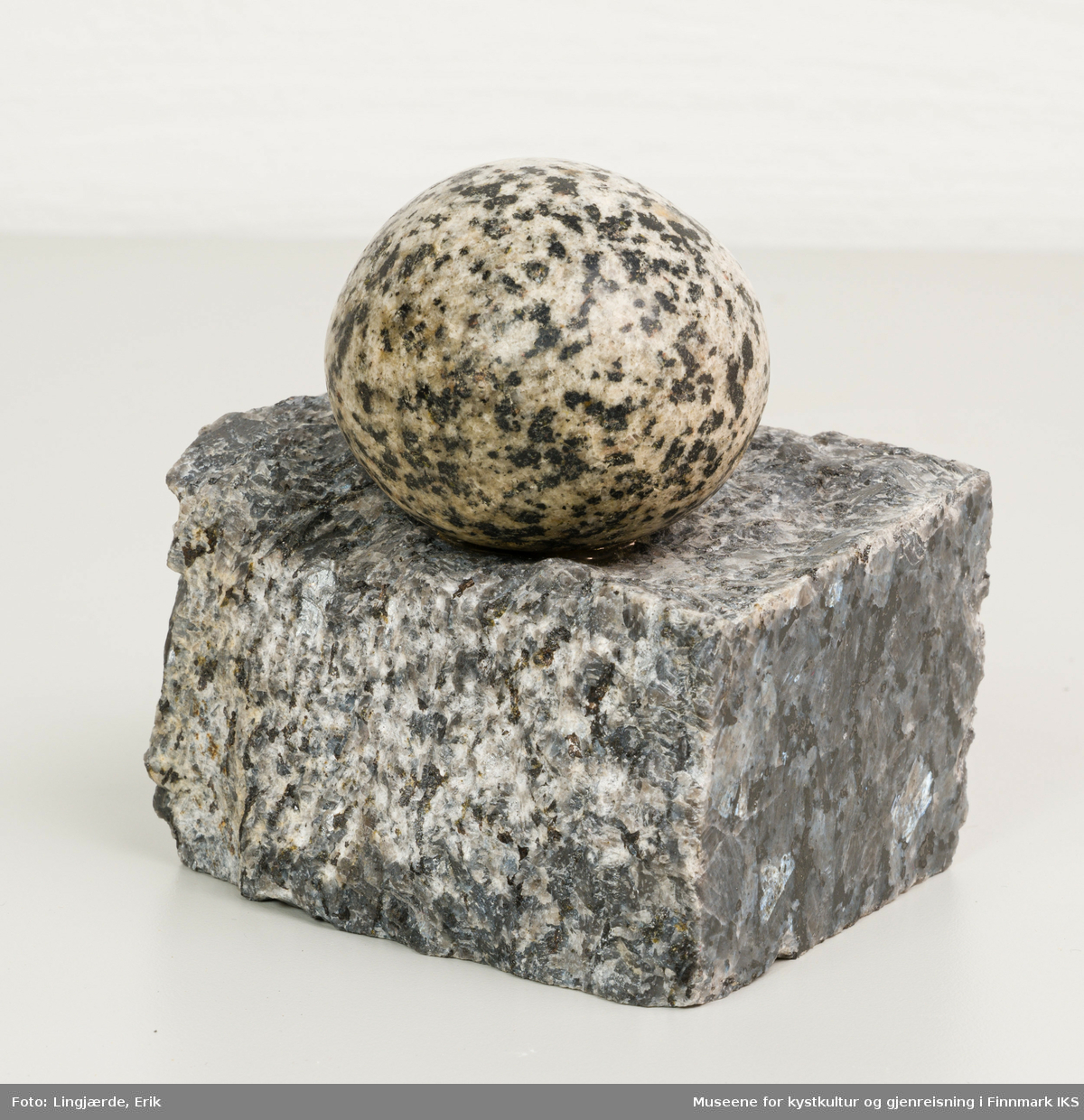 En liten oval, nesten rund stein hviler på en firkantet stein, som er glatt polert på to sider og grov på de andre to sider.