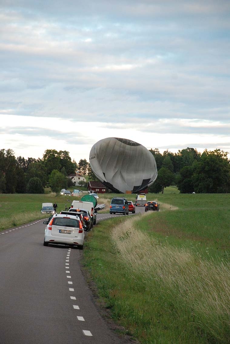 En luftballong har just landat på ett fält intill vägen mellan Adelöv och Hullaryd. Ballonghöljet håller på att lägga sig. Flera bilar parkerade utmed vägen.
