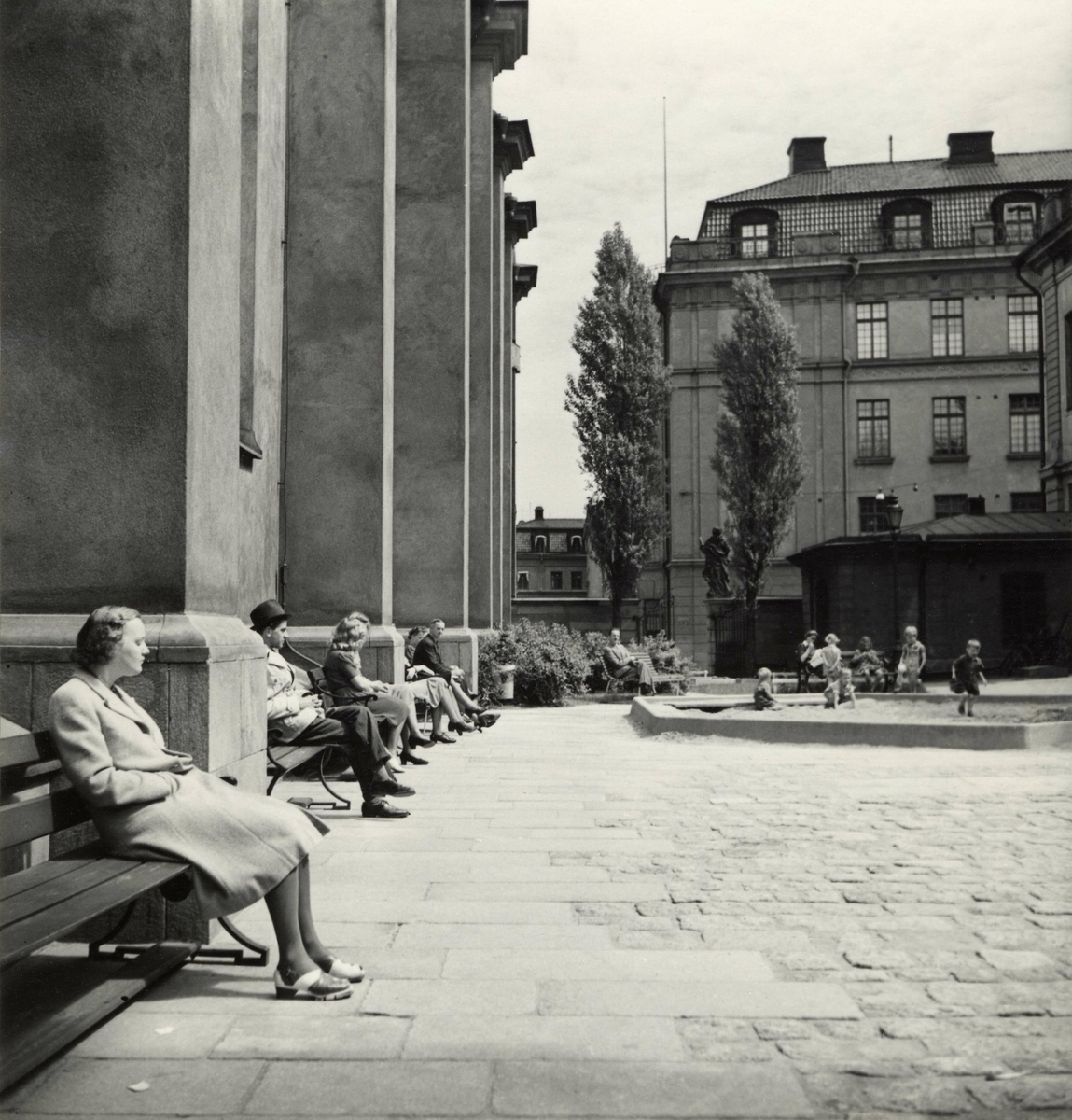 Storkyrkan, Gamla stan, Stockholm. Människor sitter på bänkarna utmed fasaden. På den öppna platsen leker barn i en sandlåda.