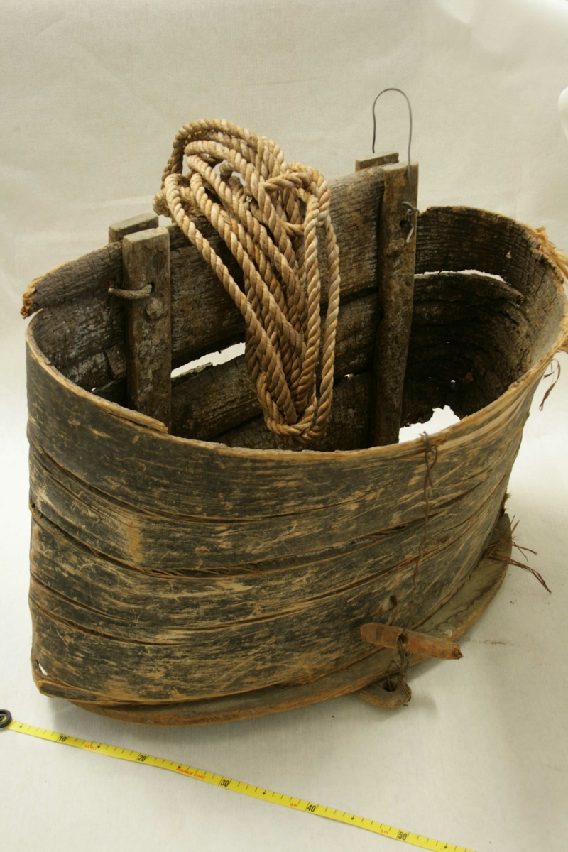 Stor laup laget av bark som er bøyd med utsiden innover, trebunn, to fester til tau, noen meter med tau ligger oppi laupen