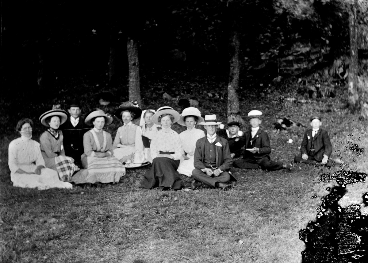 En grupp ungdomar sitter i en skogsdunge nedanför ett berg med en kaffebricka mitt i bland sig. Till höger sitter tre gossar och fotografen själv i ljus hatt, Gunnar Engström. Damerna har breda sommarhattar och möjligen är det midsommar eftersom kvinnan i mitten bär folkdräkt.