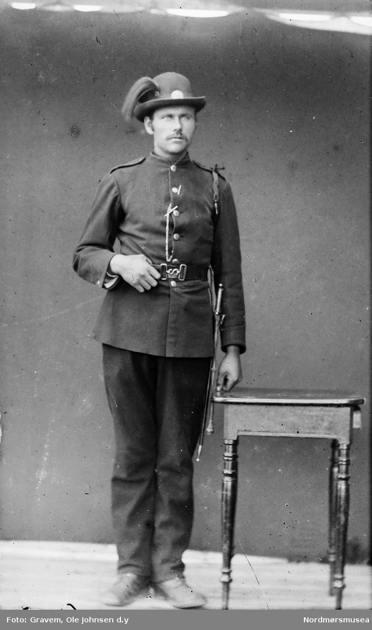 Portrett i helfigur av mann i en slags uniform. Mannen har også på seg belte og hatt.
