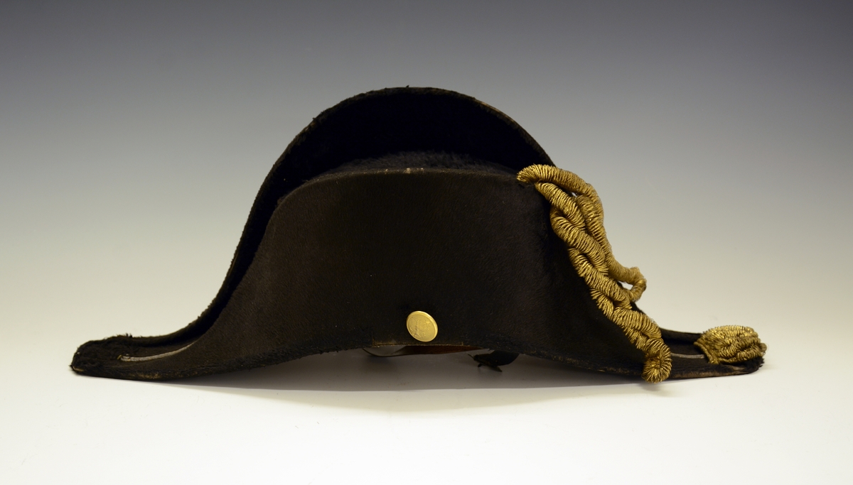 TGM-BM.1940-41:038.A Tosnutet hatt.
TGM-BM.1940-41:038.B Hatteeske
Fra protokoll:Snutehatt av filt, med vidne gullband feste til "løveknappar". Til hatten høyrer blekkøskje.

