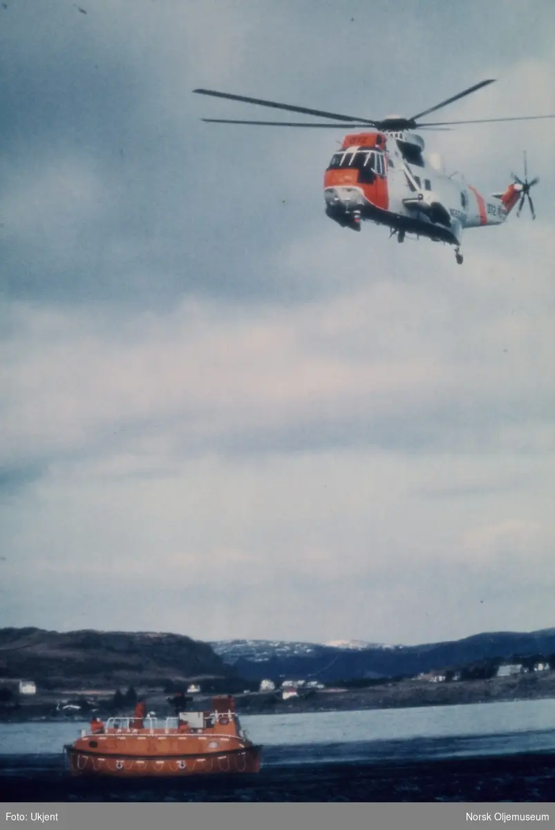 Redningsøvelse i en fjord på sørvestlandet. Et redningshelikopter sirkler over en livbåt med mannskap ombord. På bilde nr. 2 heises en livredder ned fra helikopteret.