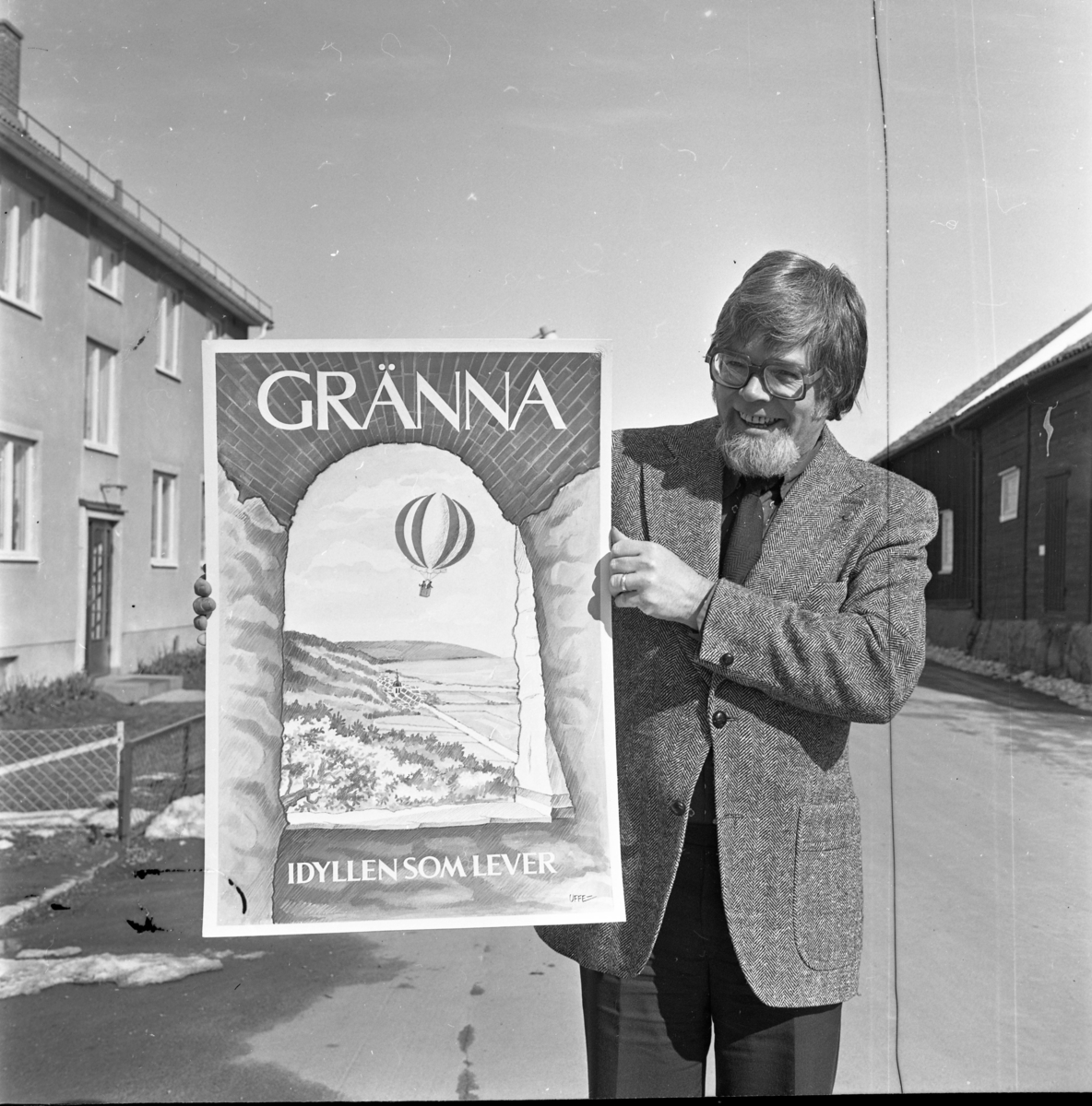 En man med skägg och glasögon håller upp en ny turistaffisch för Gränna som han har tecknat. Han står på Sjögatan i Gränna.