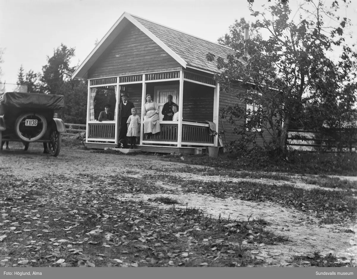 Fem vuxna personer, ett barn och en hund på verandan till en mindre stuga. Intill står Gottfrid Höglunds T-Ford 1922.