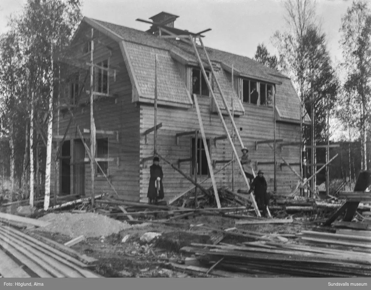 Pågående bygge av ett bostadshus i timmer med byggställningar och stege. Sex personer syns invid huset och i övervåningens fönster.