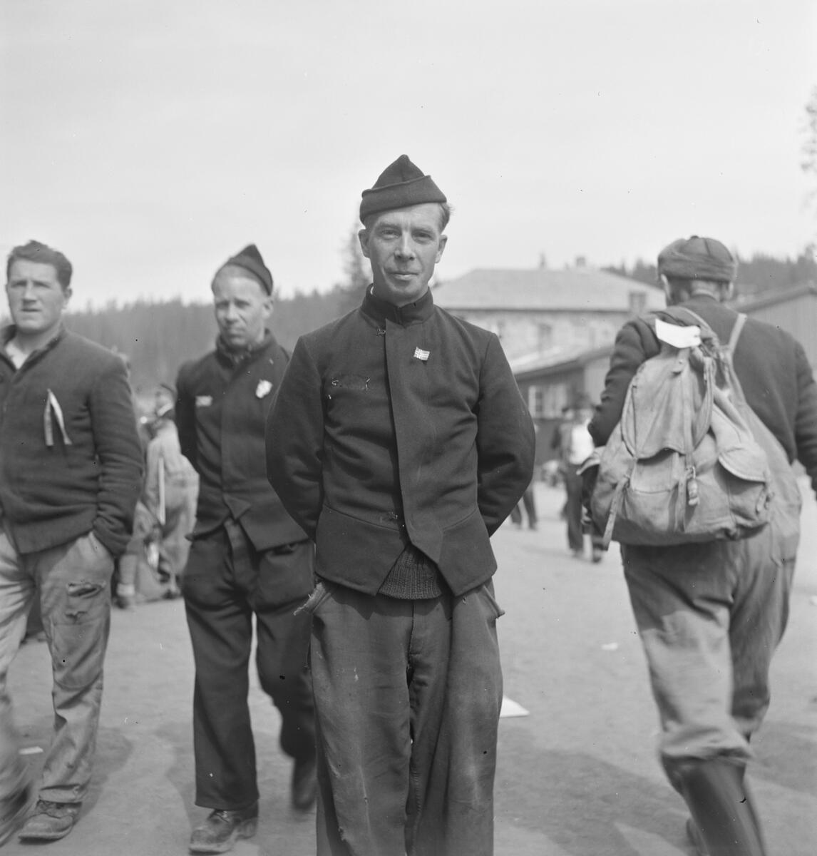 Fotografi tatt ved frigjøringen. Tre Grinifanger, med fangeuniform, er vendt med ansiktet mot fotografen. Den ene fangen står midt i bildets fokuset, med armene bak ryggen og bærer et norsk flagg i jakken sin. Til høyre for han er en person med ryggen mot fotografen. Han har en sekk på ryggen.