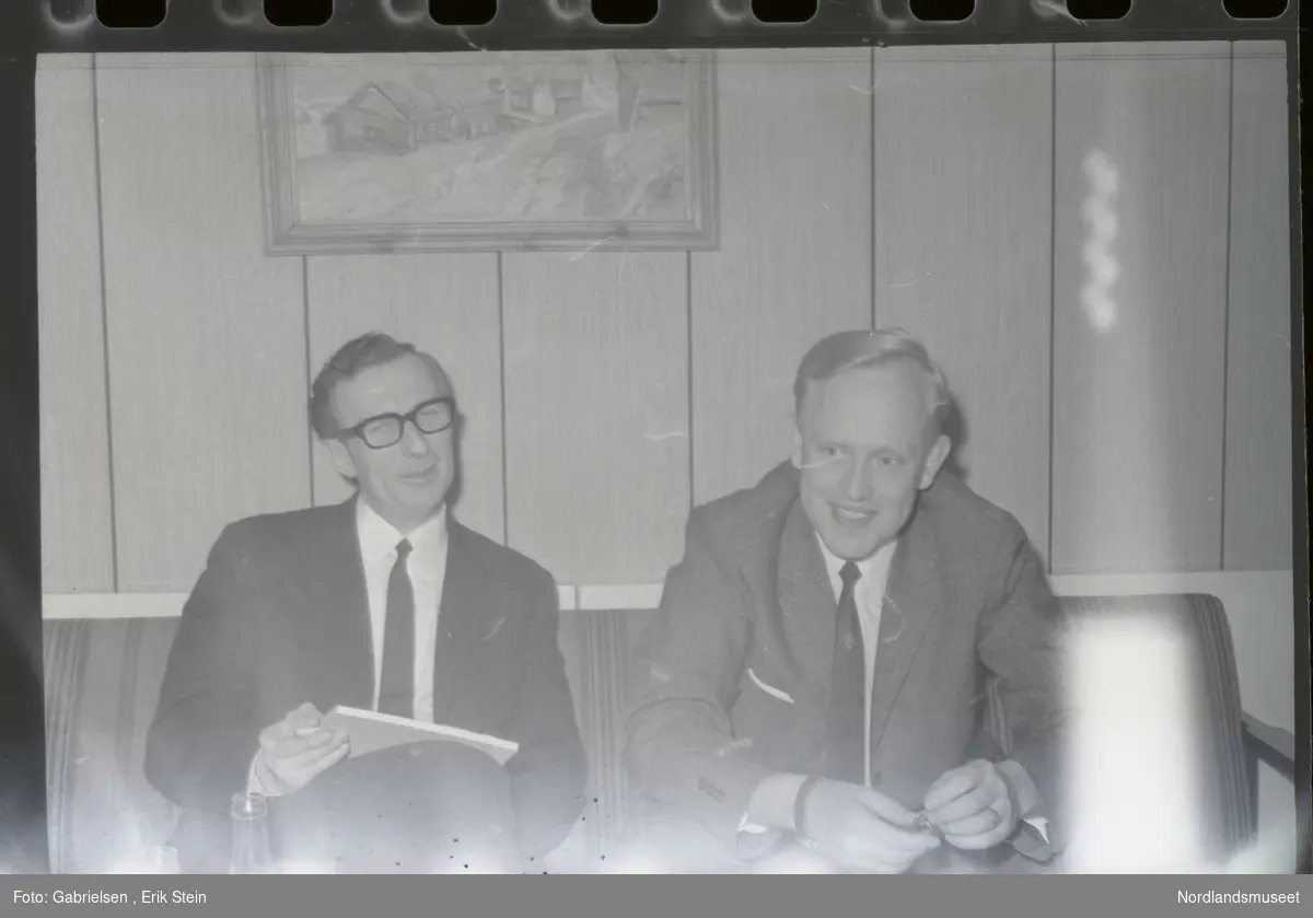 Fotografi av to menn kledd i dresser med slips som sitter
i en sofa i en stue vedsiden av en vegg som det henger et bilde på og et bord som det står noen flasker på 
og man ser at mannen til venstre med briller på seg som skriver i en papirblokk og at mannen til høyre holder
en knapp i hendene