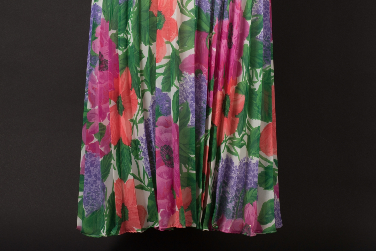 Lång skjortblusklänning i polyester med permanentplisserad kjol. 
Blommigt mönster i vitt, rosa, rött, grönt och lila. På framsidan finns tre klädda knappar och ett blixtlås. De långa ärmarna är rynkade vid handleden och knyts ihop med band. Till klänningen hör ett löst knytskärp av ett rosa och ett grönt stoppat band. 
Blixtlåset fram är fastsatt vid ett senare tillfälle samtidigt som ett knapphål och en knapp sytts dit mitt fram. 
Kjolen är fodrad med ett vitt nylontyg.

På insidans etiketter framgår att klänningen har storlek 42 och är tillverkad i Sverige för "collection philip". På tvättetiketten är tvättrådet 40 grader, ingen torktumling, låg värme på strykjärnet samt kemtvätt "P".