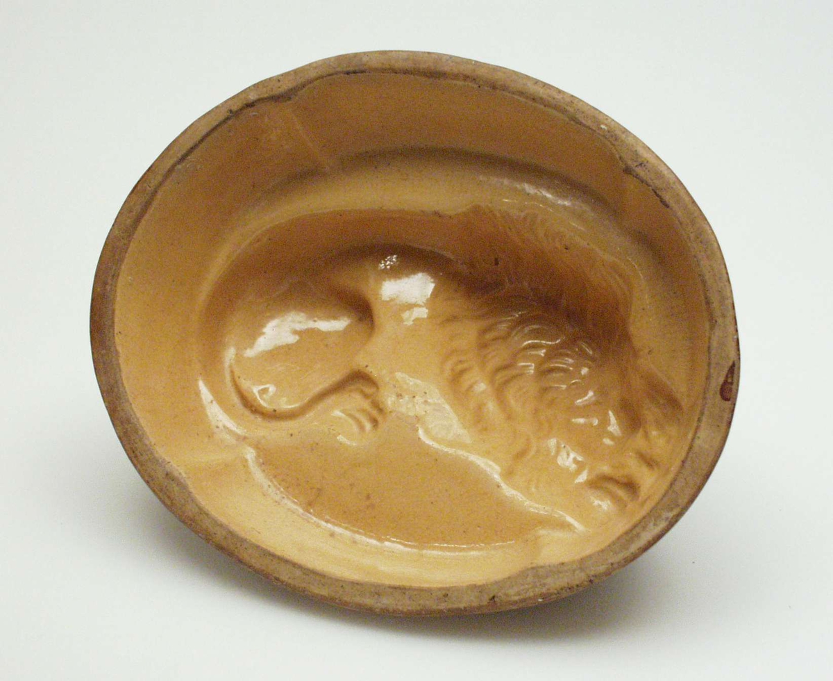 Oval puddingform i steintøy med løvemotiv. Den er brun utenpå og gul inni.