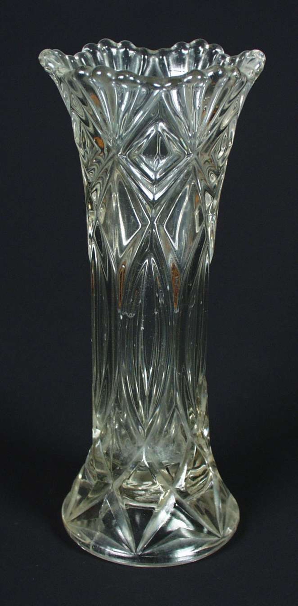 Vase i glass med sylindrisk form, utvidet mot fot og topp. V-formede linjer i kryss, linjeform. Vasen har bølgeformet kant øverst med kuleformede topper innimellom, seksdelt. Foten er sirkelrund og innhul.