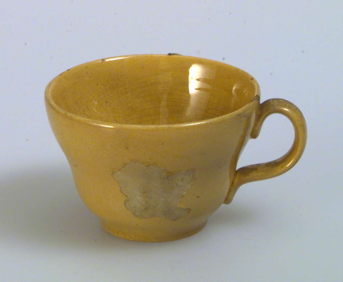 Gul kaffe-/tekopp med skål. Dekoren på koppen er falt av. Skålen er uten dekor. Det er hakk i både koppen og skålen.
Det er ingen produksjonsmerker på kopp eller skål.