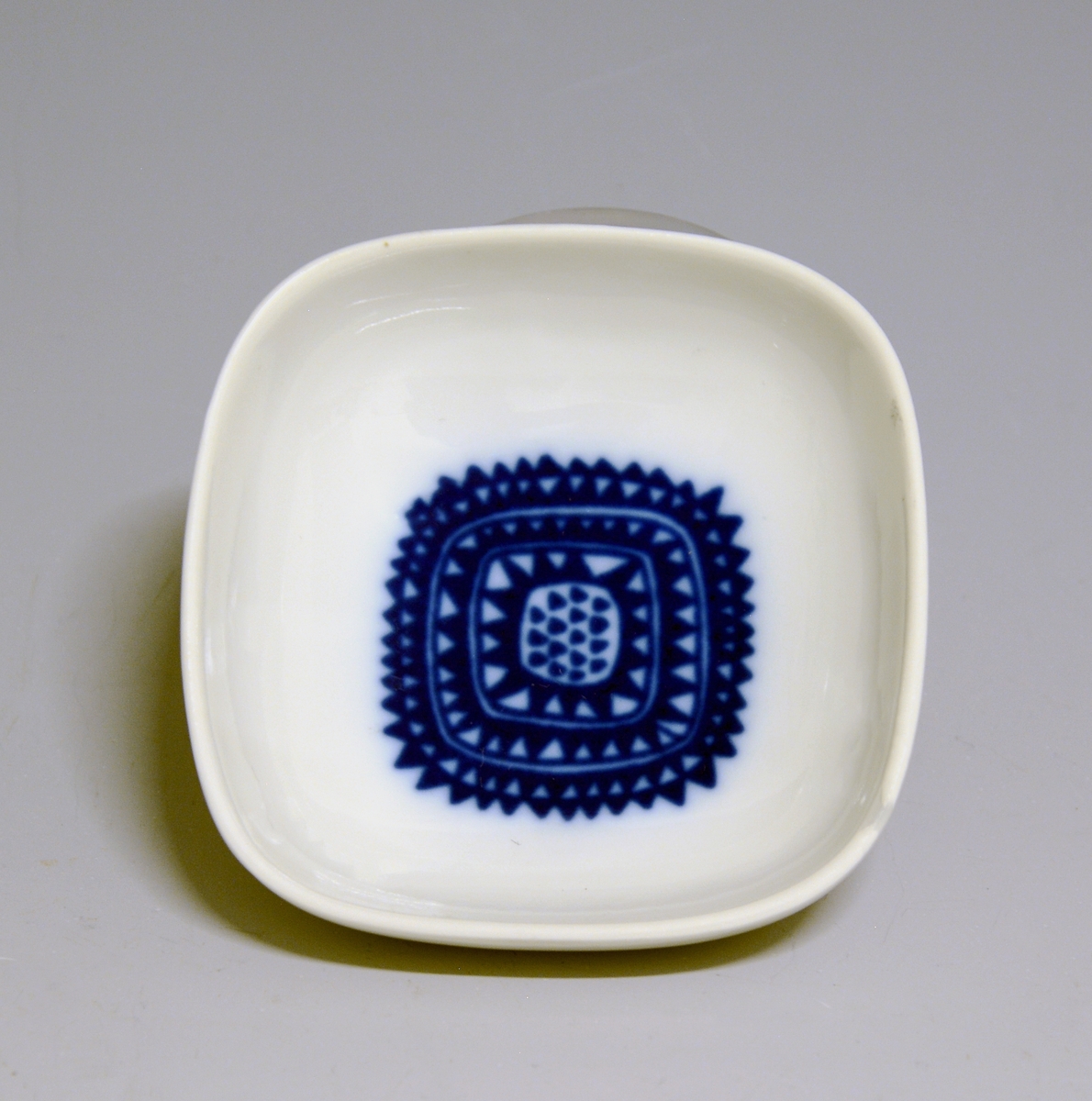 Prot: Askeskål av porselen. Samme som TGM-BM.1968:324 men dekoren er geometrisk mønster i mørk blå farge. Mrk: PP 67 og M.2259, D. 78107. Design Konrad Galaaen, dekor: Arne Lindaas.