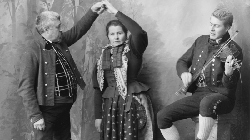 Dette er et gruppebilde der mann og kvinne ikledd Rørosdraktdanser pols. Felespiller sitter ved siden av. (Foto/Photo)