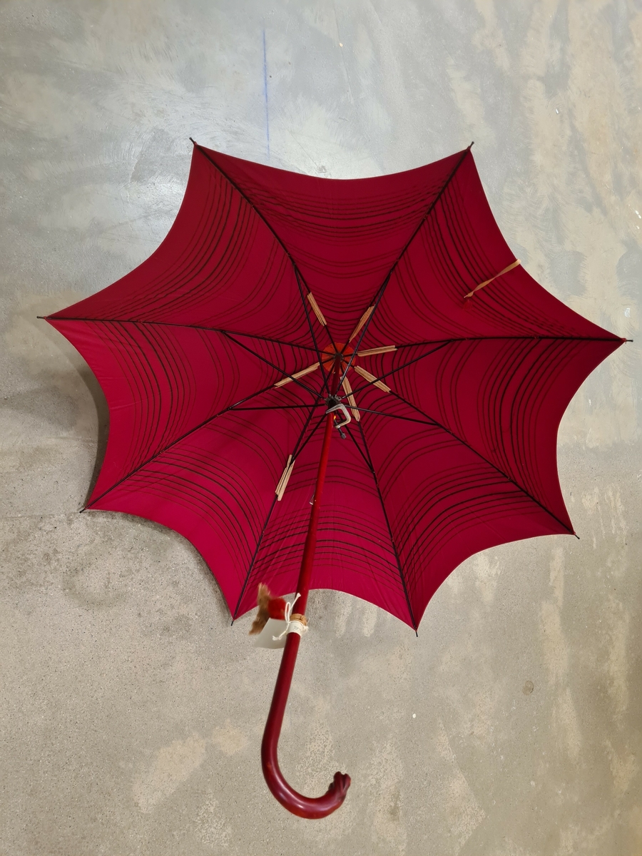 Paraply i rött tyg med svart randning. Krycka som är klädd med rött läder. Vid kryckan hänger två tofsar.

Ingår i en samling av nio olika parasoller med olika utseende och modell.