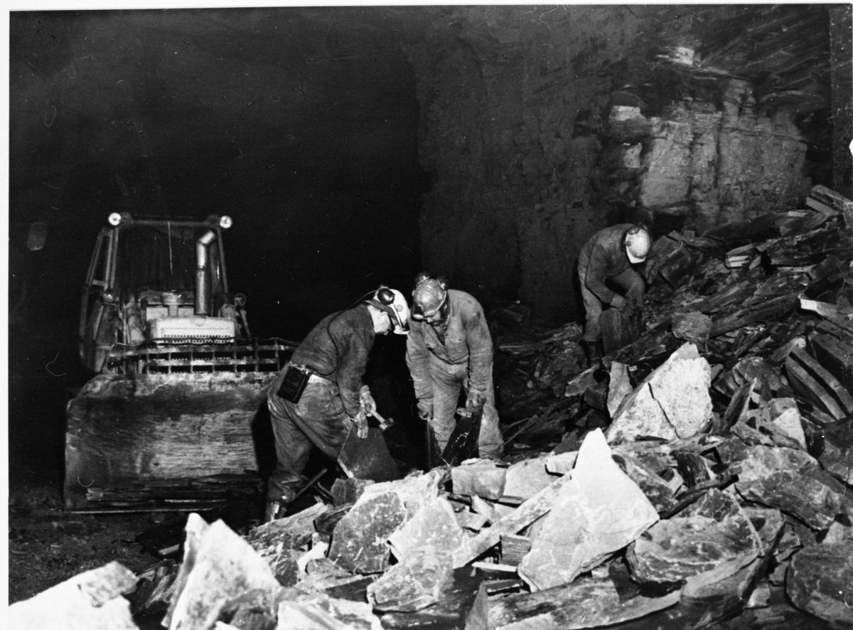 Tre menn arbeider ved en av gruvene til Valdres Skiferbrud. Mennnen er iført arbeidsklær og vernehjelm.