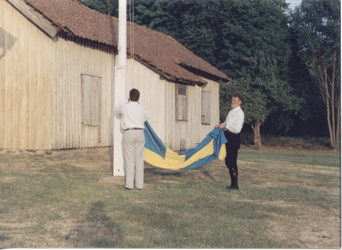 Staffan Bjerrhede och Berny Gustafsson hissar flaggan inför hembygdsgårdens midsommarfirande 1993. Hembygdsgården Långåker 1:3 ligger till vänster (ej i bild) och i bakgrunden ses Långåker 1:2 "Davids", "Derras".
Relaterade motiv: A03389 - A03408.