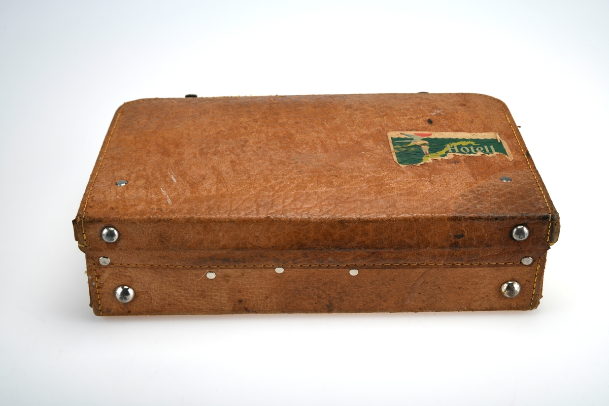 Koffert laget av lær, stoff og pappkartong med tilhørende nøkler. 

Alle hjørnene både på bunn og lokk er forsterket med påsydd skinn. På toppen av kofferten er det et håndtak. De er festet med stift på innsiden. Det er to koffertlåser, en på hver side av håndtaket. På bunnen av kofferten og under har den fire metallnagler som fungerer som ben slik at den kan stå eller ligge uten å bli skadet. På innsiden av kofferten er det rosa/beige stoff samt flere lummer og bånd for å holde gjenstander på plass. Fire lommer i lær på innsiden av lokket og åtte bånd i lær i bunnen av kofferten. Lommene i lokket inneholder en saks, en fil og et reisespeil, den siste lommen er tom men ser ut til å ha kunnet hylstre en kam. Lommen med reisespeilet har et enkelt feste i lær for å holde speilet på plass. 

Ellers inneholder kofferten 3 glassbeholdere i ulike størrelser med lokk, 2 børster, 1 sammenleggbar flaskeåpner, 1 tube med "Pond's cold cream", 1 eske som tidligere har inneholdt avføringsmiddel "Relaxit" og 1 plastbeholder som inneholder 2 uåpnede såper med ulik embalasje fra tyske Mäurer & Wirtz.