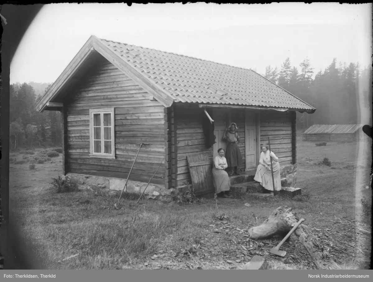 Tre kvinner foran laftet bygg på Tovestul. To av kvinnene har redskap i hånden. Øks liggende ved stubbe foran huset. Rive støttet opp til veggen.