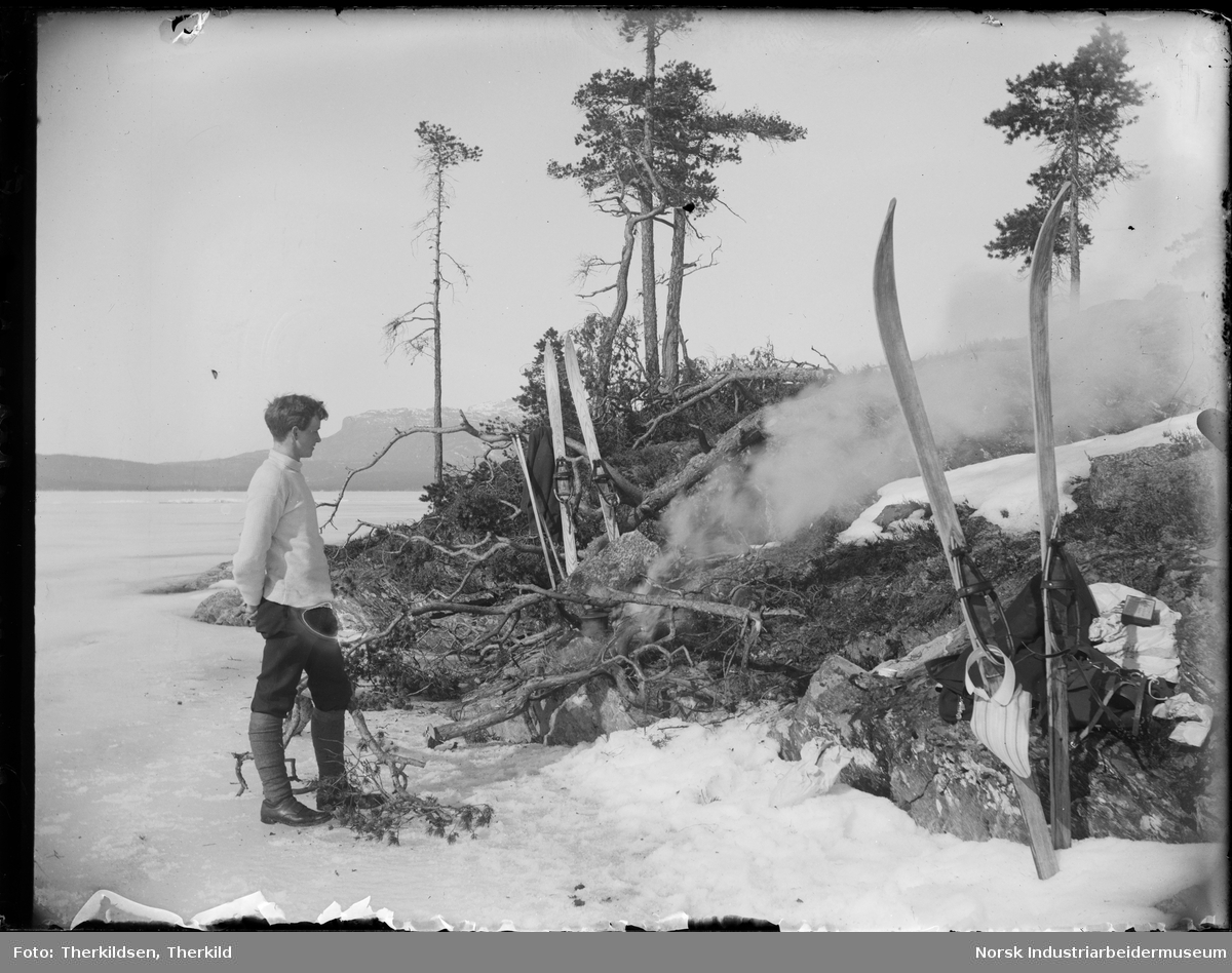 Mann stående og ser på bål. Et par ski på hver side av bålet. Mannen står på islagt vann, antagelig Follsjå.