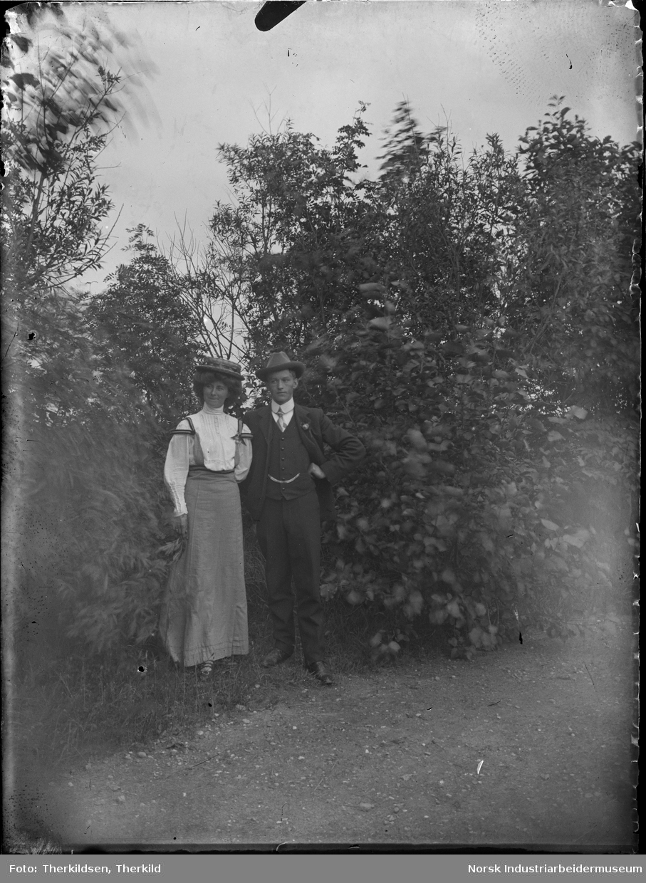 Kvinne og mann i pentøy oppstilt foran busker og trær.