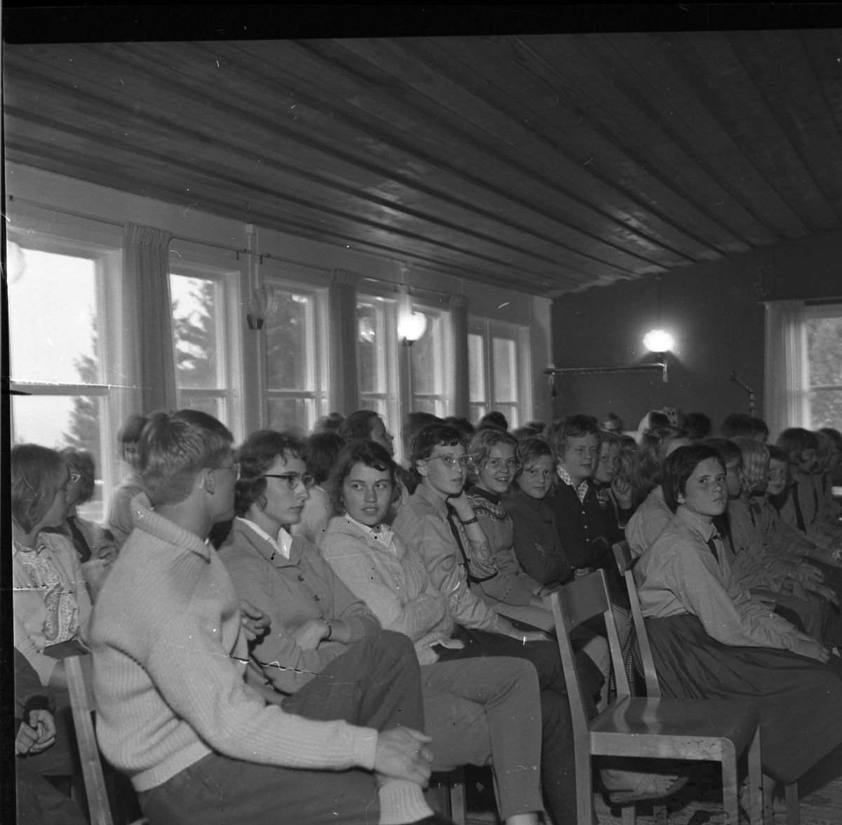 Kvinnor och män och några barn sitter och lyssnar på en talare som inte syns i bild. Vissa tittar mot kameran.