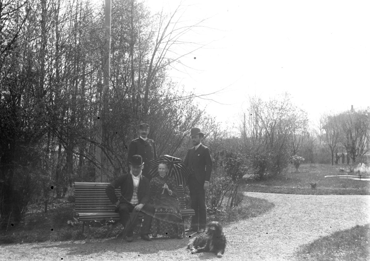 Tre menn med hatt og en kvinne med parasoll samt en hund på/v benk i park med springvann.