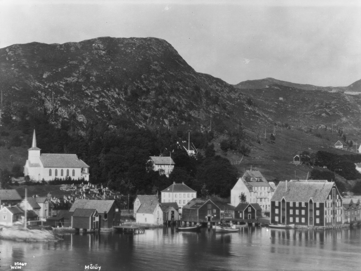 Måløy med hus og lagerbygning i sjøkanten, kirken med kirkegården og fjellet i bakgrunnen.