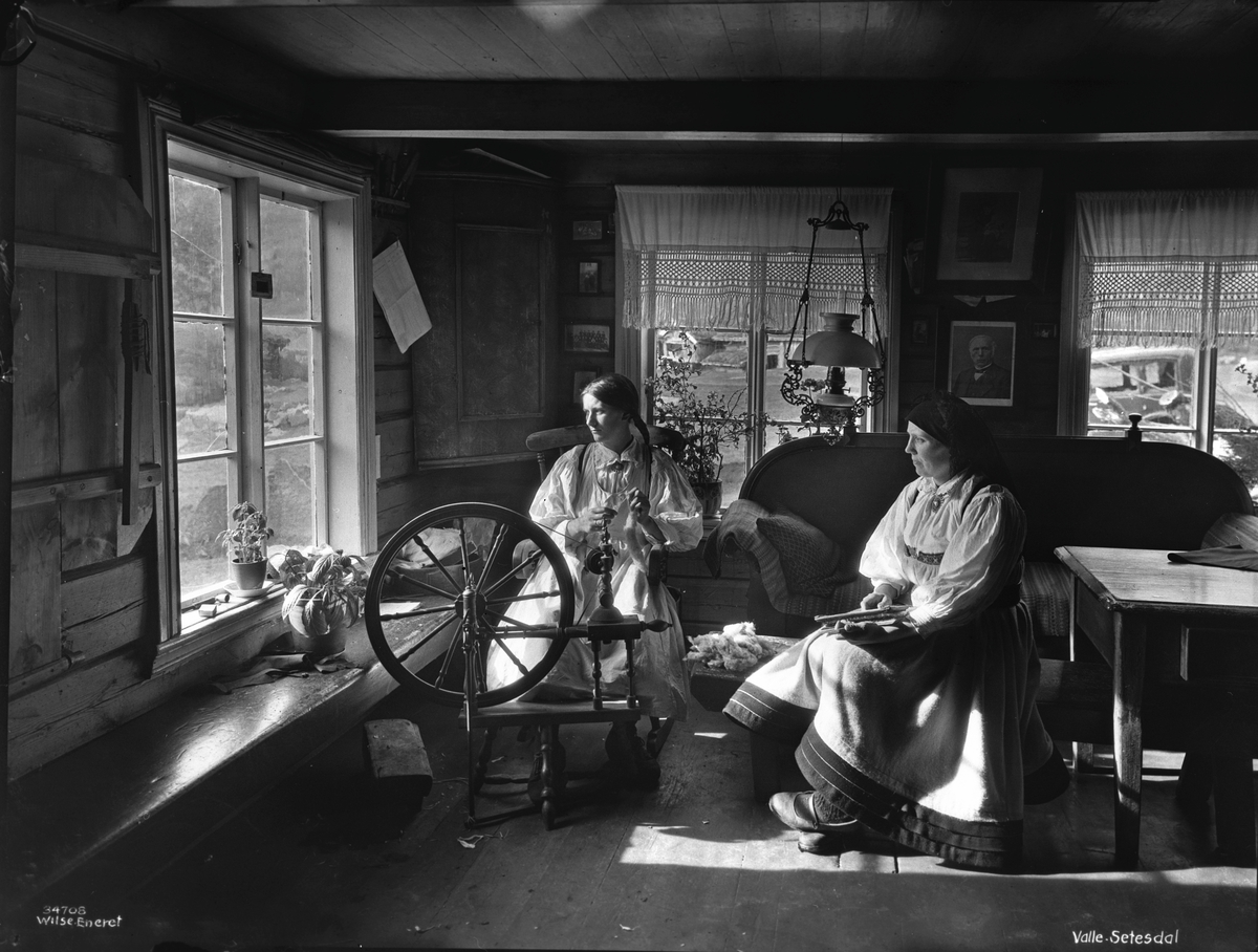 To unge kvinner i Setesdalsdrakt sitter ved rokken, lyset skinner inn i stuen fra de tre vinduene.