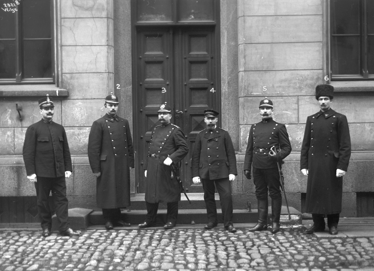Seks politikonstabler i uniform avbildet utenfor inngangen til bygning, antatt politistasjon.