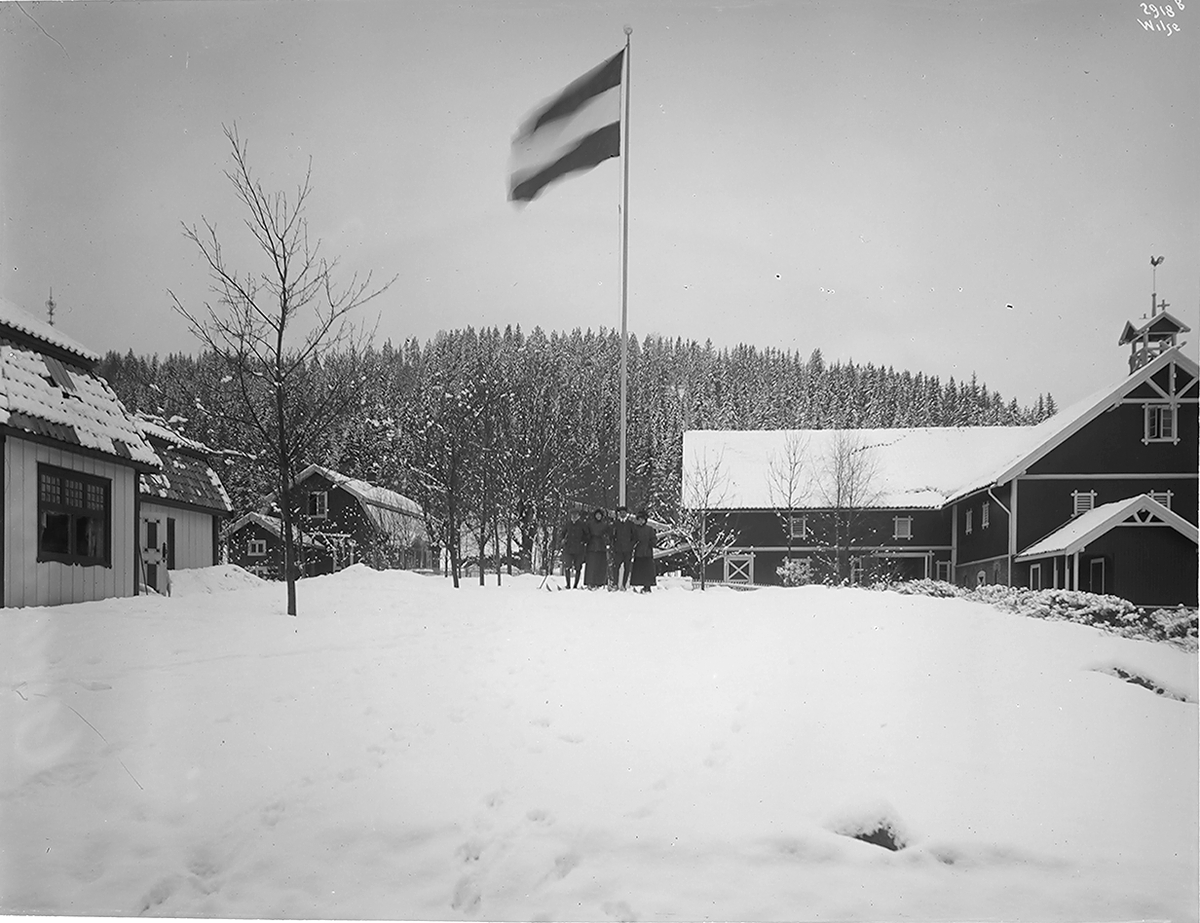 Godseier Ribberts villa, her avbildet gårdsplassen med fjøs og låvebygning. Familien Ribbert fotografert ved flaggstangen.