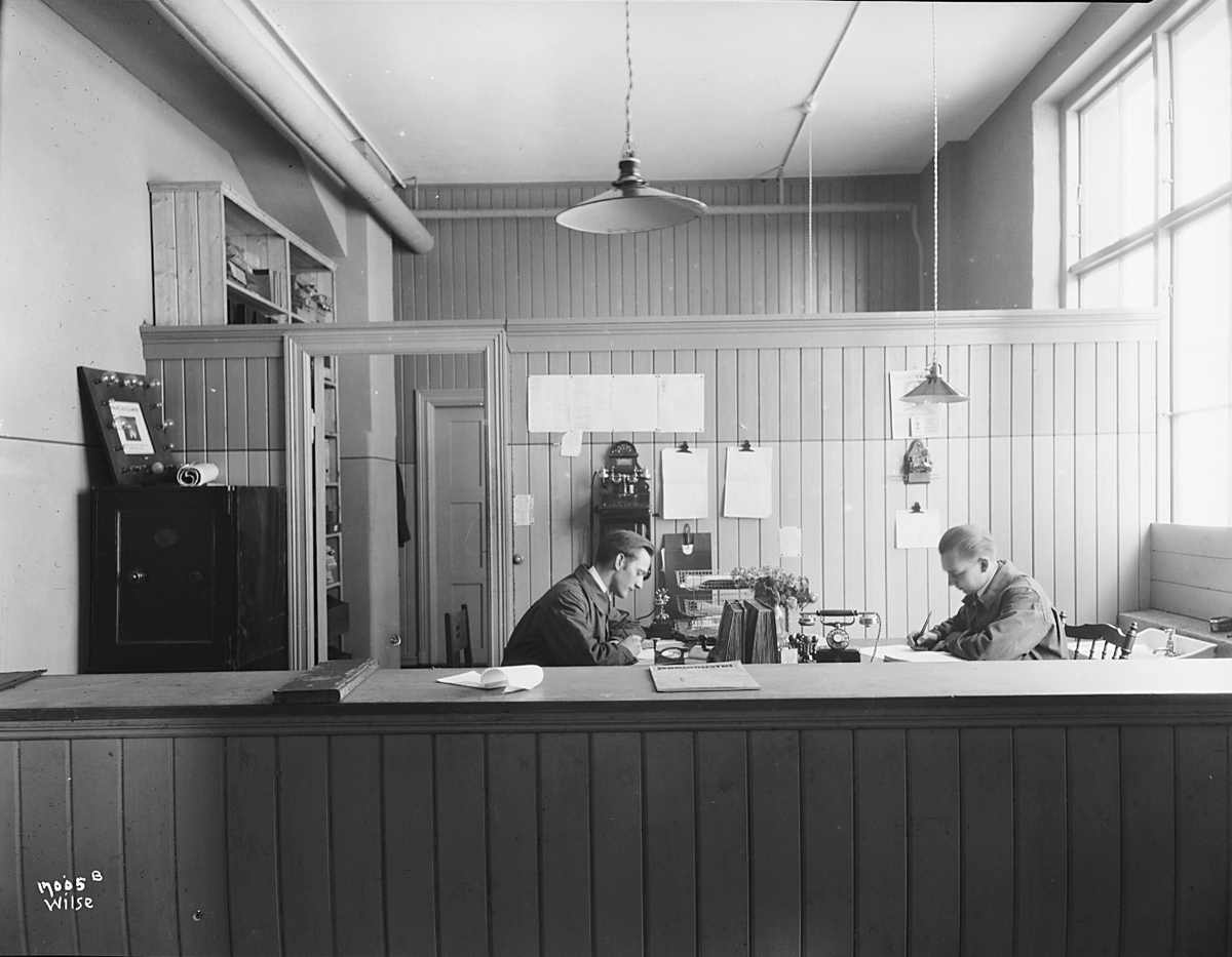 To kontorister sitter ved skrivebord bak en skranke, Sørensen og Balchen A/S. Fotografert 1923.