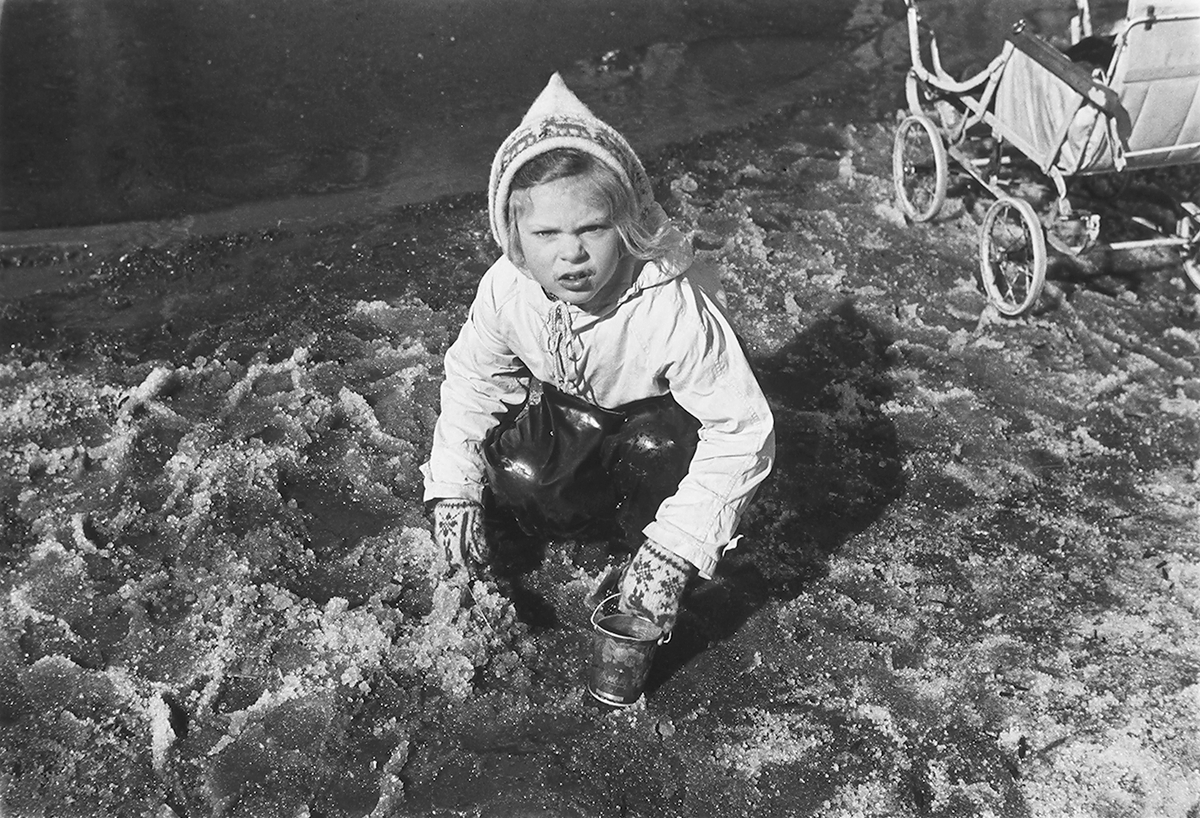 Pike med bøtte og spade i snøslaps. Barnevogn i bakgrunn. Fotografert 1940.