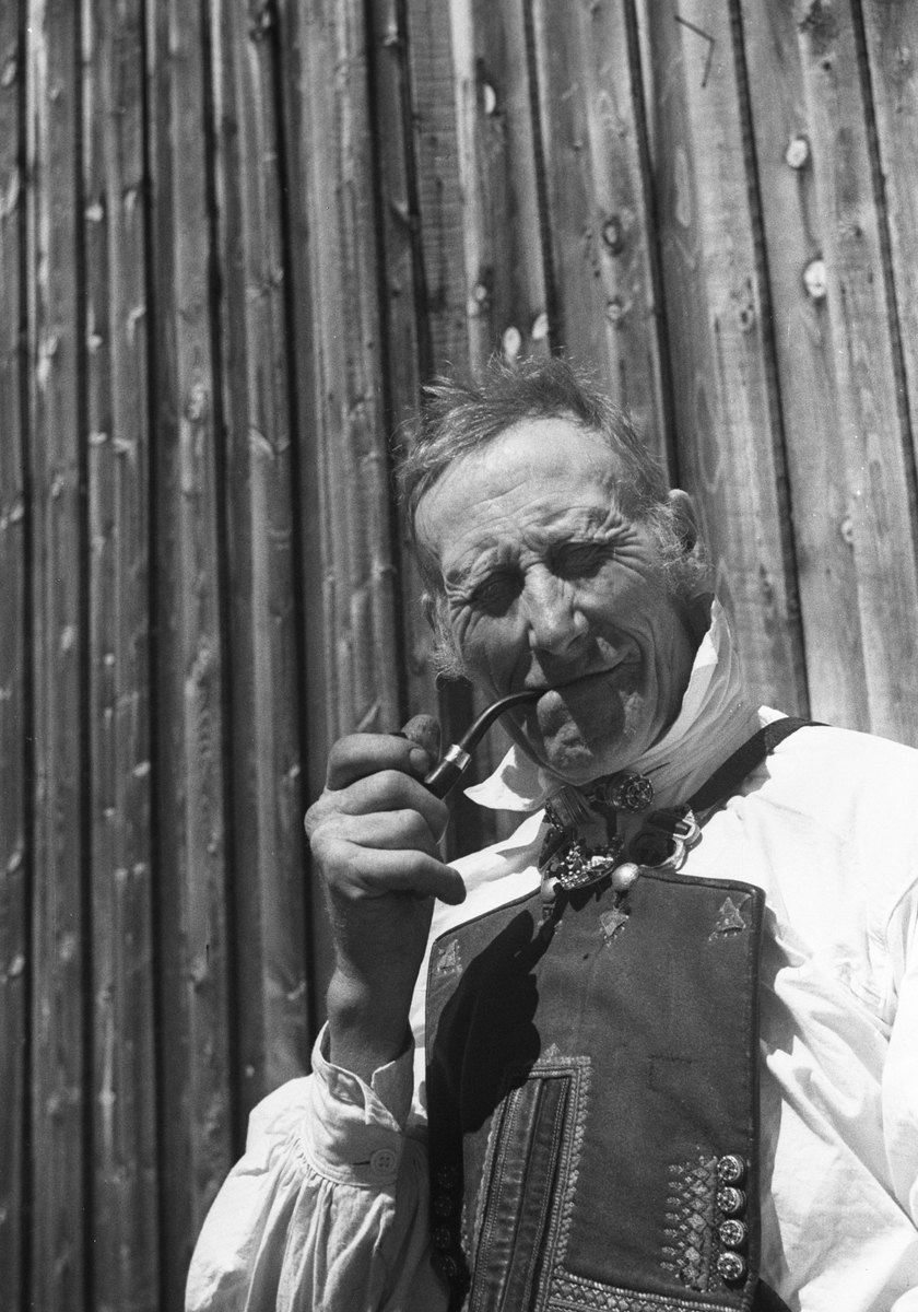 Mann i Setesdalsbunad med pipe i hånden. Sitter i solskinnet ved uthusveggen.