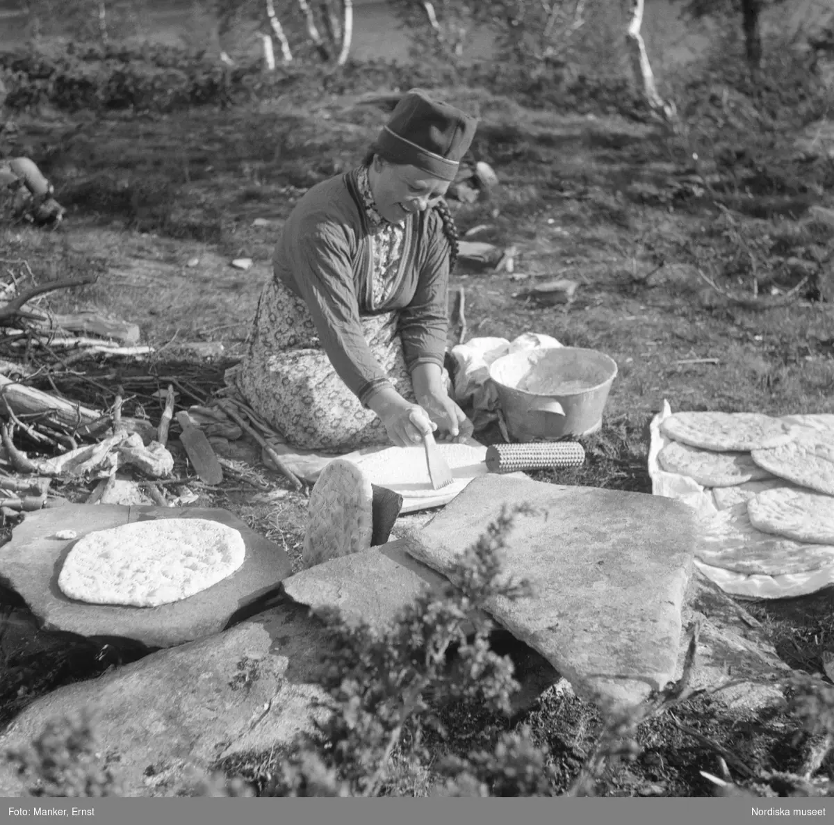 En kvinna sitter och bakar tunnbröd utomhus, det är Maria Pavval som gräddar bröd på en stenhäll utanför kåtan.