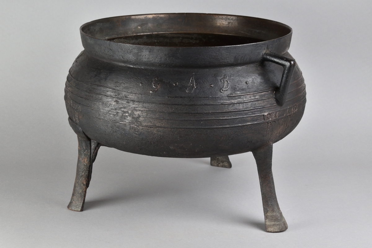Gryta tillverkad i malm. Rund till formen med utåtvinklad kant, två vinklade öron och tre fötter. Buken med upphöjda ränder samt stämplad "Anno 1695", "G.J.D" och "E.A.D".
