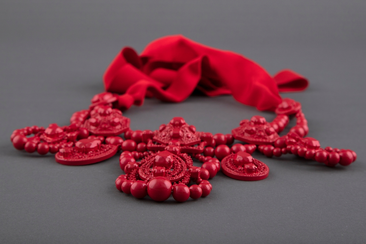 Stort halssmykke av rødlakkert metall, plastperler og brede tekstilbånd i samme farge. Smykket knytes i nakken.