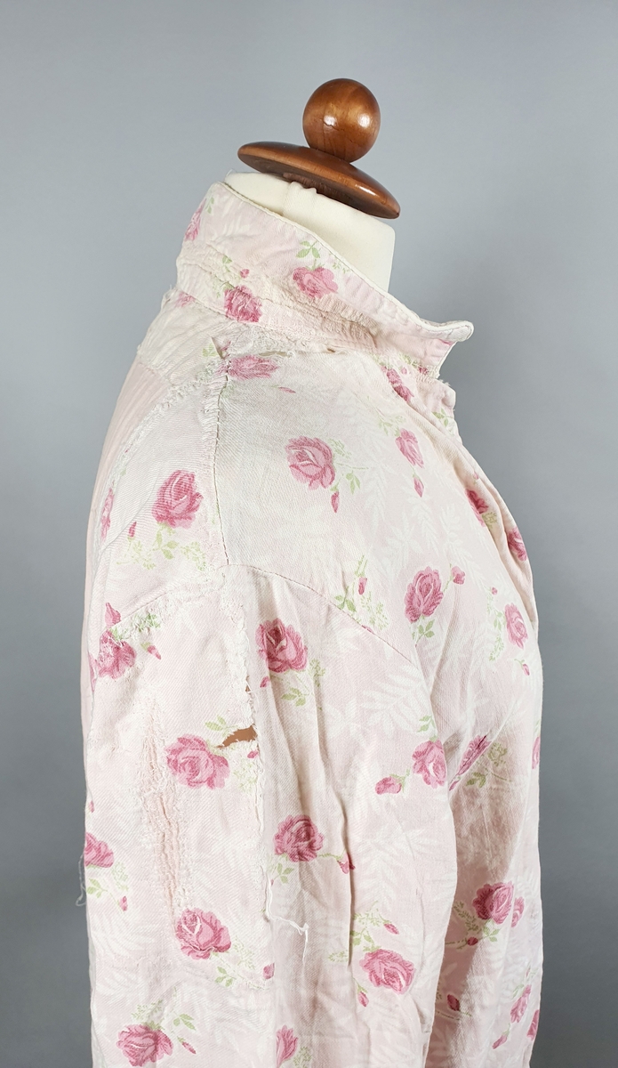 Rosa nattskjorte av bomull med rosemønster. Skjorten har ulike knapper i front, og er lappet i nakke og på ryggen og er slitt (delvis revnet) på skudre og øverst på ermene. Skjorten er forlenget nederst med rosa bomullstoff.