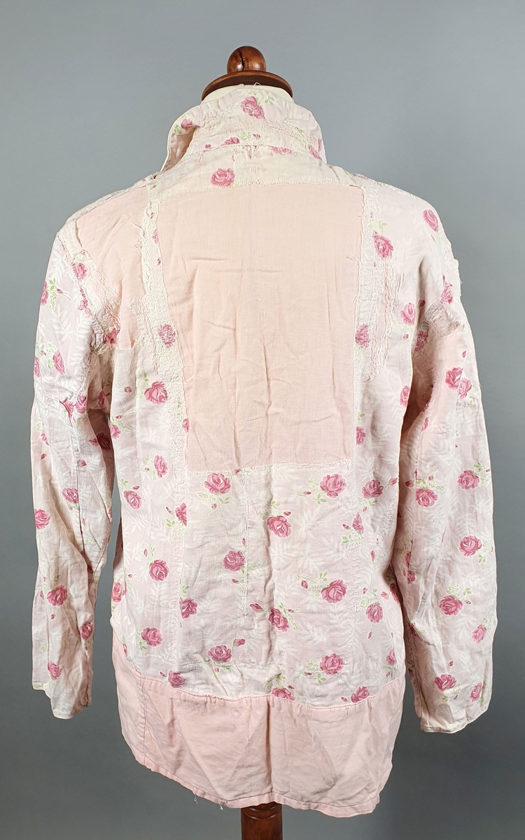 Rosa nattskjorte av bomull med rosemønster. Skjorten har ulike knapper i front, og er lappet i nakke og på ryggen og er slitt (delvis revnet) på skudre og øverst på ermene. Skjorten er forlenget nederst med rosa bomullstoff.