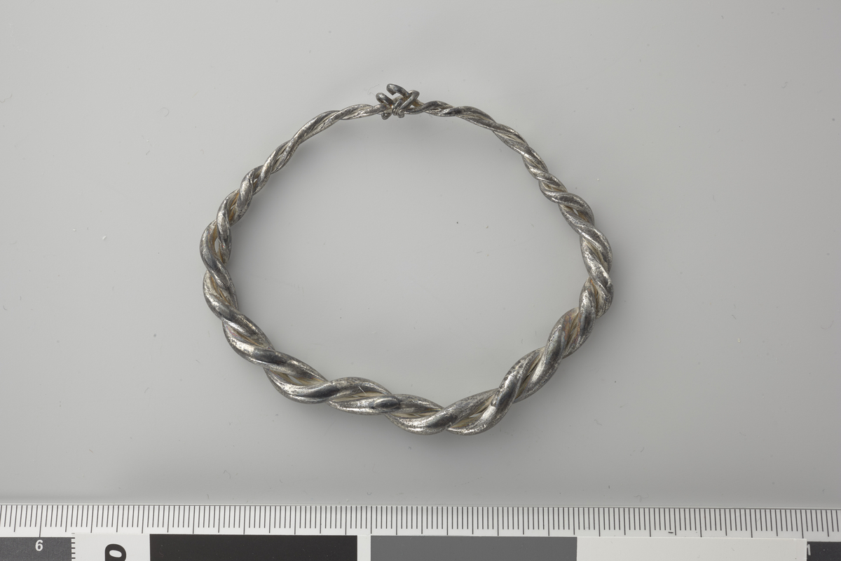  Ganske liten håndleddsring av sølv dannet av to sammensnodde sølvtener, sl. Grieg: Vikingetidens Skattefund, s. 255, fig. 54 øverst til venstre. Ringens tykkelse er størst på midten, den avtar jevnt mot endene, hvis tener er ganske enkelt sammensnodd. Ringen er nu litt forbøiet. Største tverrmål 8,3 cm, største tykkelse 1 cm. 