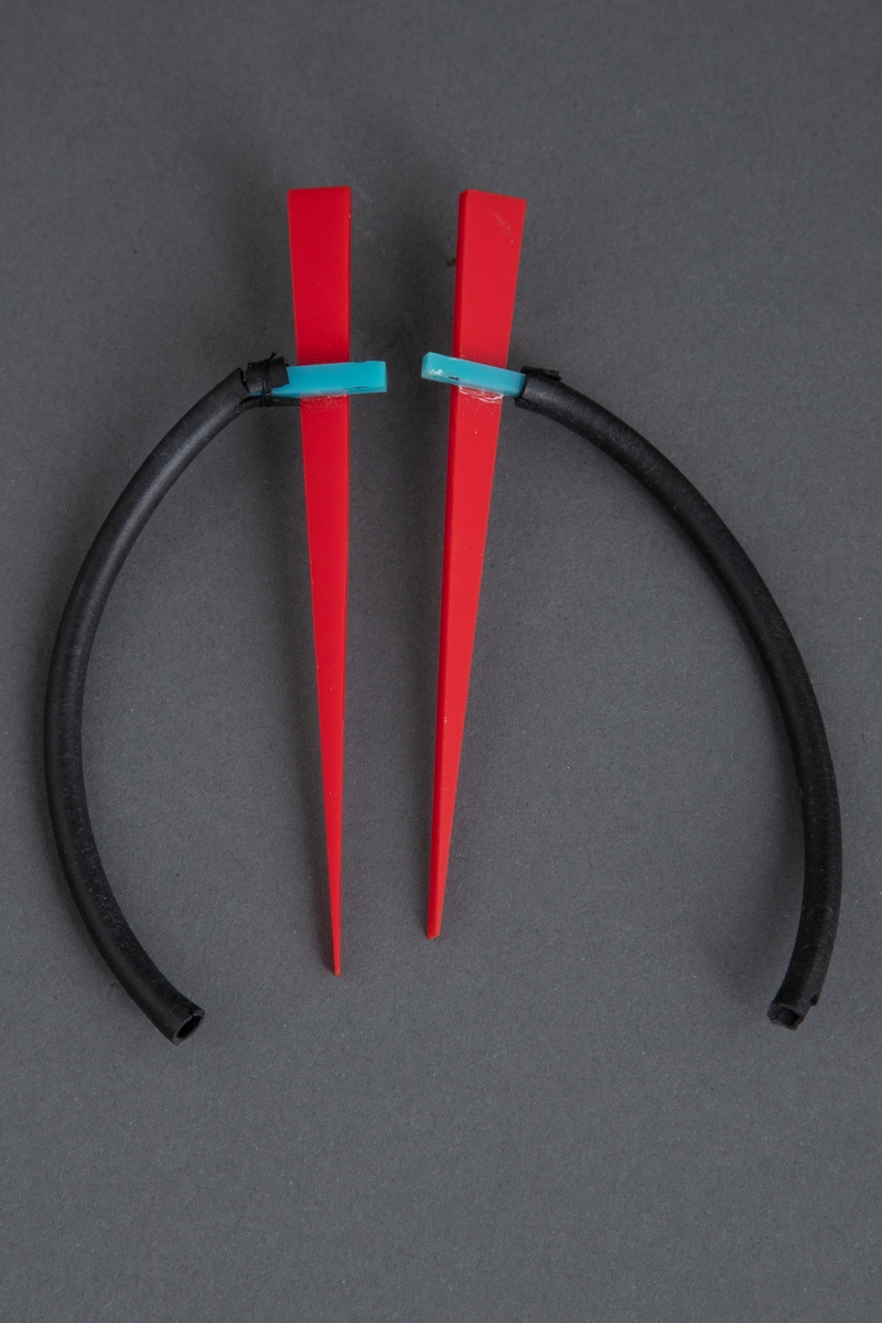 Et par nesten identiske øresmykker som består av en spiss trekant i rød plast. På denne er det pålimt en lys blå plastbit. Til denne biten er det festet en svart gummislange, slik at den nærmest danner en sirkel. På baksiden av den røde plasten er det pålimt en stift for å feste øresmykket til øret.