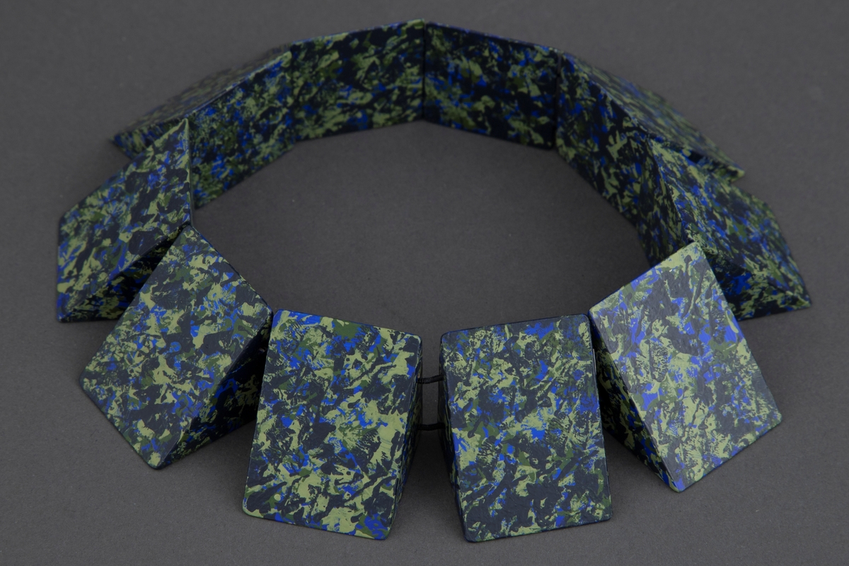 Halssmykke bestående av trestykker med form som en halv kube som er tredd på en dobbelt, svart hattestrikk. Trestykkene er malt i svarte, skarp blå, lyse- og mørkegrønne sjatteringer.