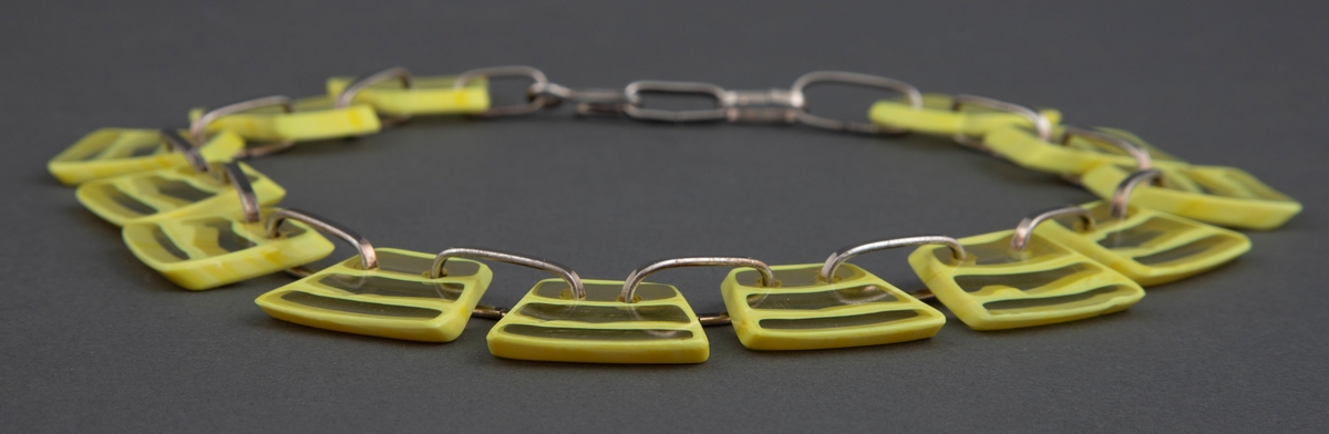 Halssmykke med store, trapesformede glassbiter i gult og klart glass, der klare og gule horisontale striper er omkranset av gult. Glassbitene er hengslet sammen med grove sølvringer.