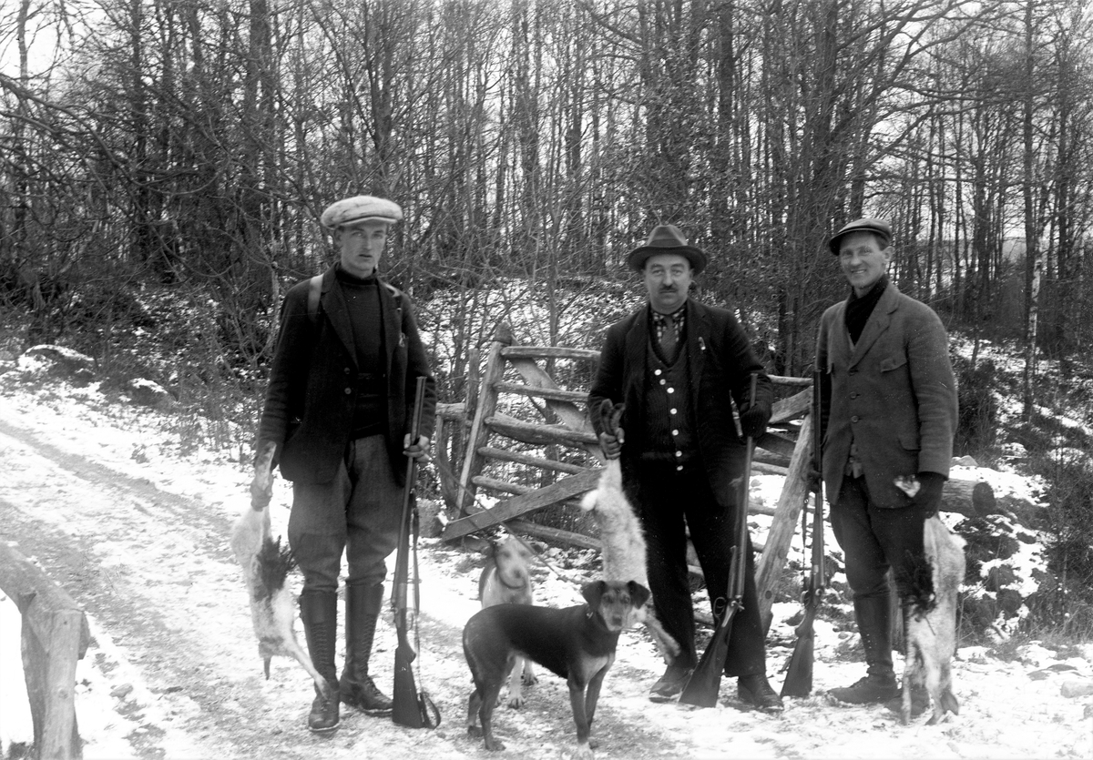 Tre jägare står med gevär, skjutna harar och ett par jakthundar på en smal väg vid en öppen grind. Marken täcks av ett tunt snötäcke. Risknippen har placerats i hararnas uppskurna magar.