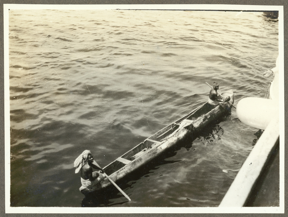 Bilden föreställer två svarta män som kör förbi Fylgia i en stockbåt.
