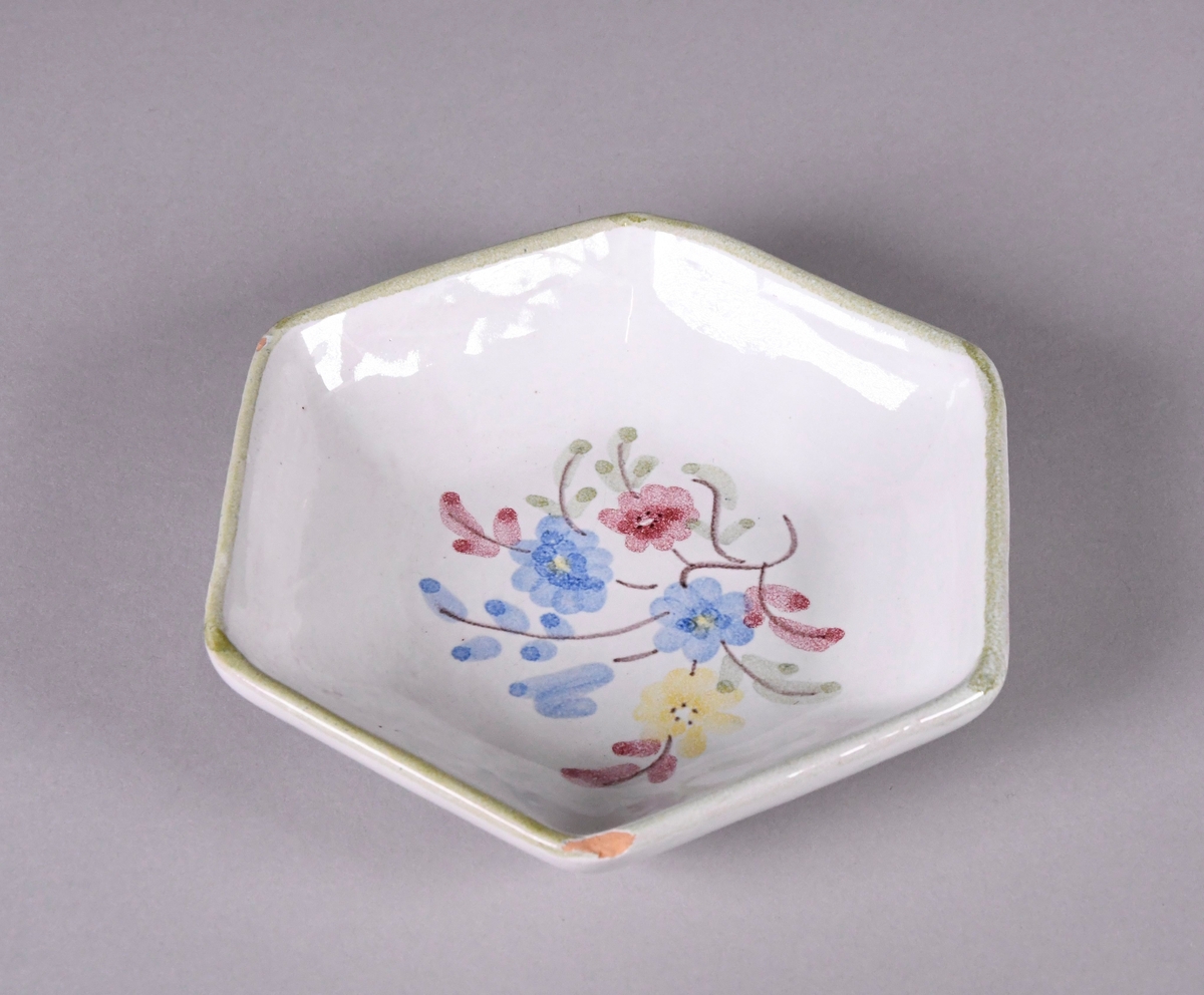 Skål av glassert keramikk, med opphøyd rand. Skålen er sekskantet, og har hvit grunnfarge. Håndmalt dekor av røde gule og blå blomster og en grønn borde langs randen.